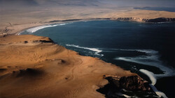 Aerial view of a vast desert coastline meeting a deep blue ocean with white surf patterns.