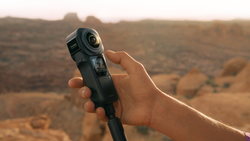 Hand holding a compact 360-degree action camera against a desert landscape backdrop.