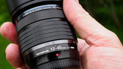 Hand holding an Olympus 12-45mm f3.5-5.6 telephoto zoom lens against a blurred green background.