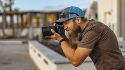Bearded photographer in blue cap and brown shirt shooting with a DSLR camera on an urban rooftop.