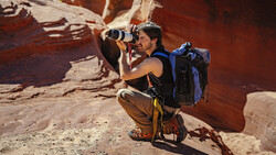 Adventure photographer crouching in desert canyon while photographing with telephoto lens and backpack.