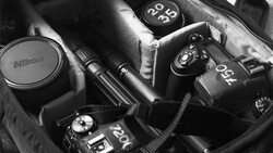 Collection of Nikon DSLR cameras and lenses arranged in a camera bag, shot in black and white.