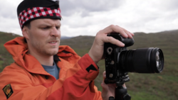 Photographer in orange jacket and red plaid headband holding a DSLR camera with telephoto lens in an open moorland landscape.