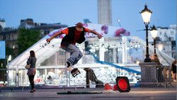 Skateboarder performing a trick mid-air above a rail with an urban fountain and cityscape in the background.