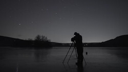 Silhouetted photographer on frozen lake at night with telescope on tripod pointed at starry sky.