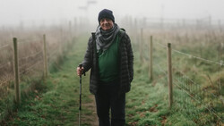 Person standing in a foggy vineyard row holding a walking stick, wearing winter clothing and a beanie.