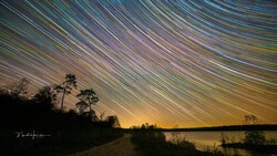 Star trail arcs sweep across the night sky above a dark lakeside landscape with silhouetted trees.