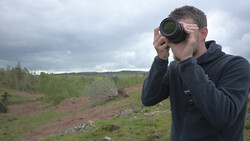 Photographer holding binoculars to his eyes while standing in a rolling landscape.