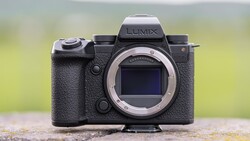 Panasonic Lumix mirrorless camera body with lens mount visible, positioned on a textured surface with blurred green background.