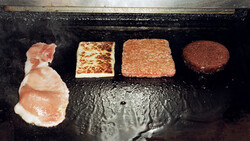 Four food items at different cooking stages on a dark griddle: raw prosciutto, lightly cooked patty, medium-cooked patty, and fully cooked dark patty.