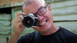 Smiling photographer holding a silver mirrorless camera with a wide-angle lens up to his face.