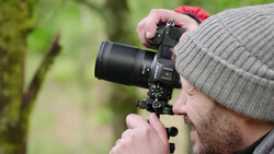 Photographer holding a DSLR camera with telephoto lens while looking through viewfinder outdoors.