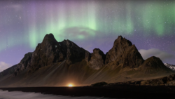 Jagged volcanic mountains silhouetted against a green and purple aurora borealis display over dark water.