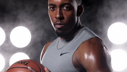 Athletic man in sleeveless gray shirt holding basketball against dark background with dramatic lighting.