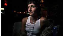 Person in white tank top and plaid shirt against dark background with warm ambient lights.