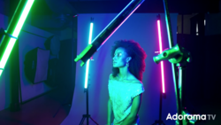 Woman posed among neon tube lights in cyan, green, pink, and blue creating a geometric light setup.