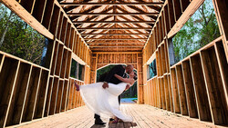 Bride in white dress twirling inside wooden barn structure with open sides and geometric roof framing.