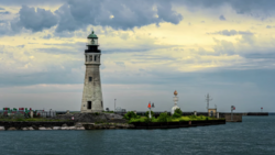 Historic lighthouse with stone tower on rocky island surrounded by water under dramatic cloudy sky.