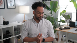 Man in light gray button-up shirt gesturing while speaking in a bright home office with plants and shelving visible behind him.