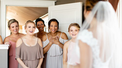 Bride in white dress viewed from behind as bridesmaids in pastel dresses smile at camera.