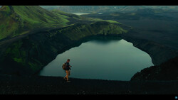 Hiker in red jacket standing on dark volcanic terrain overlooking a turquoise crater lake surrounded by green mountains.