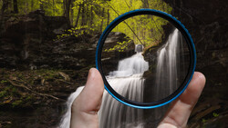 Hand holding blue circular filter frame against cascading forest waterfall.