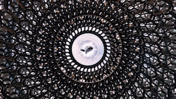 Looking up through a geometric lattice structure at a circular opening framing the sky and birds in flight.