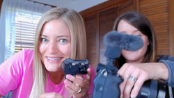 Two women holding cameras indoors, one in pink shirt holding a compact camera, the other in dark clothing holding a DSLR with windscreen attachment.
