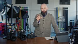 Male photographer pointing at camera in studio surrounded by professional equipment and lighting gear.