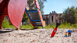 Children playing on a sandy ground with large red and blue sculptural structures and brick buildings in the background.