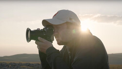 Videographer holding a cinema camera against backlit golden hour sky with hills in distance.
