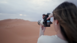 Photographer holding a mirrorless camera with telephoto lens while shooting in a desert landscape.