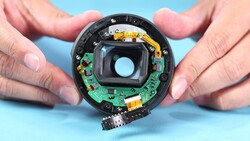 Disassembled camera lens showing internal circuit board, springs, and optical elements held in hands against blue background.