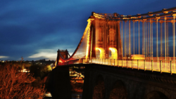Suspension bridge illuminated in golden light at dusk against a dramatic blue sky.