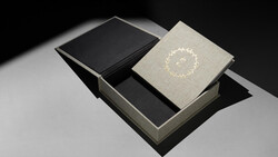 Luxury book packaging with a cream linen-covered volume featuring a decorative wreath design, displayed with a black book and gray box.