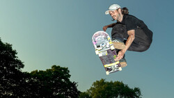 Skateboarder airborne against clear sky with trees below, wearing white cap and black clothing.
