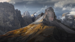 Dramatic mountain landscape with layered peaks and golden-lit slopes beneath stormy clouds.