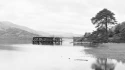 Wooden boathouse structure reflected in still water with forested shoreline and mountains beyond.