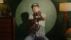 Man in pinstriped shirt and cap pointing directly at camera against large green circular backdrop.