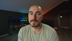 Man with shaved head and goatee wearing light gray v-neck t-shirt in dimly lit room.