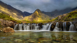 Cascading waterfall flows across layered rock formations into an emerald pool, with a distinctive peaked mountain rising beyond under dramatic cloudy skies.