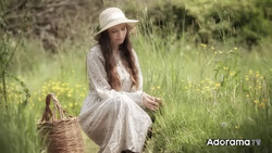 Woman in white dress and hat crouching in wildflower meadow beside wicker basket.