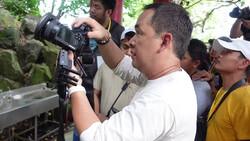 Videographer operating a cinema camera with follow focus system at an outdoor location surrounded by crew members.