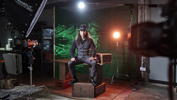 Photographer sitting on wooden box in studio with professional lighting setup and green backdrop.