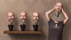 Man holding oversized head sculpture while three identical head sculptures sit on wooden shelf against patterned wallpaper.