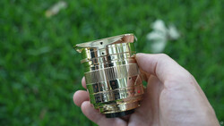 Hand holding a polished brass lens barrel with ribbed detailing and a wooden focus ring against blurred green foliage.