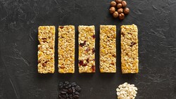 Five granola bars arranged in a row on dark textured surface with ingredient clusters displayed below.