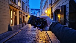 Photographer's hand holding a DSLR camera pointed toward a cobblestone street in a European city at dusk.