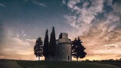 Cypress trees flanking a historic stone chapel at dusk beneath a starlit sky.