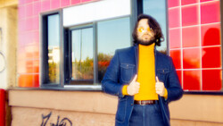 Person in denim jacket and yellow shirt standing against pink tiled wall with windows.
